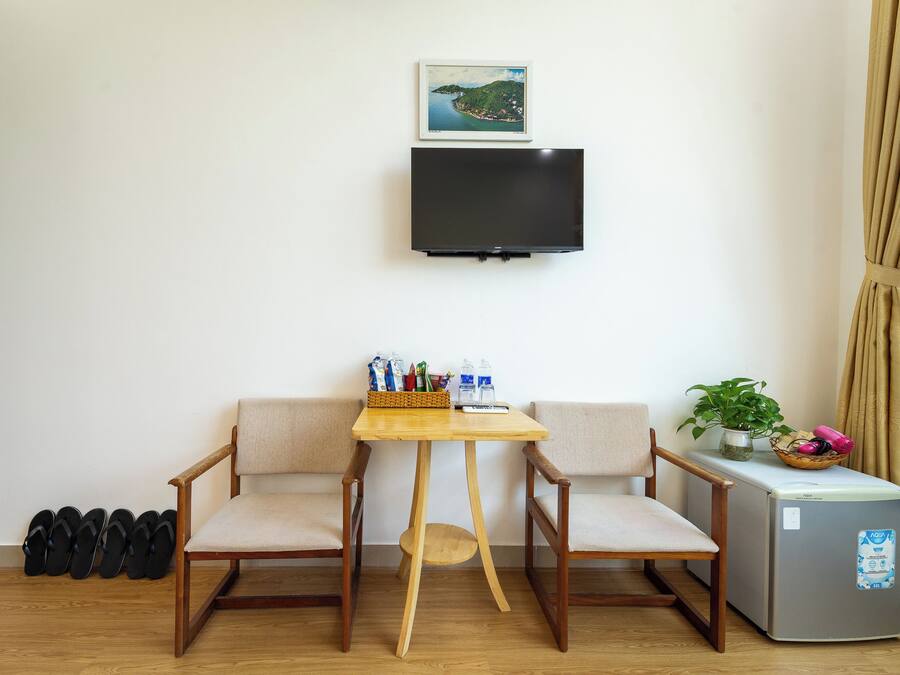 Ein kleiner Raum mit an der Wand montiertem Fernseher, einem Holztisch mit Stühlen, einem kleinen Kühlschrank und einem Paar Slippers an der Wand.