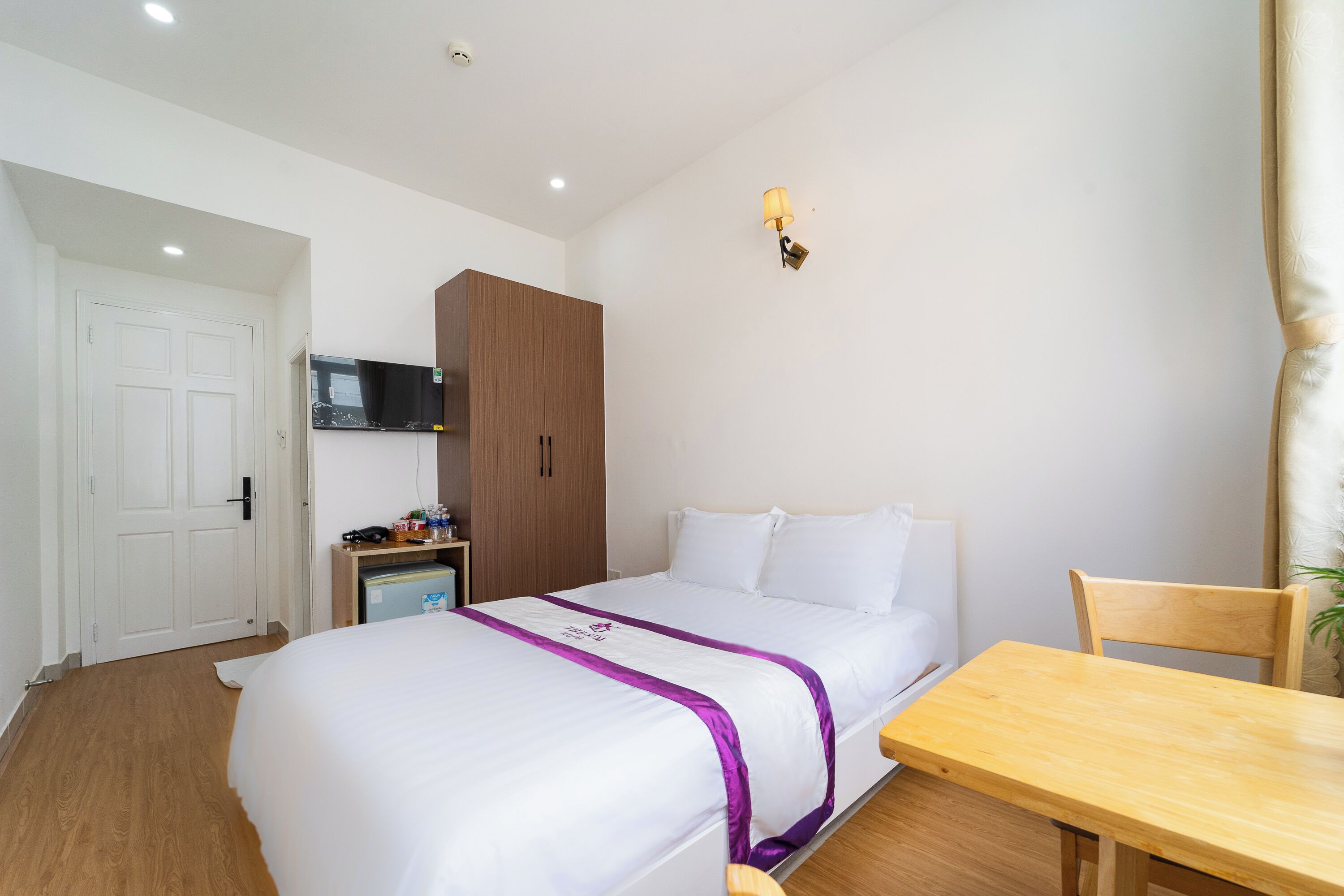 A hotel room with a bed, a wooden table, a TV, a wardrobe, and a lamp.