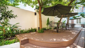 Een houten terras met buitenmeubilair, omgeven door groen en een witte muur.
