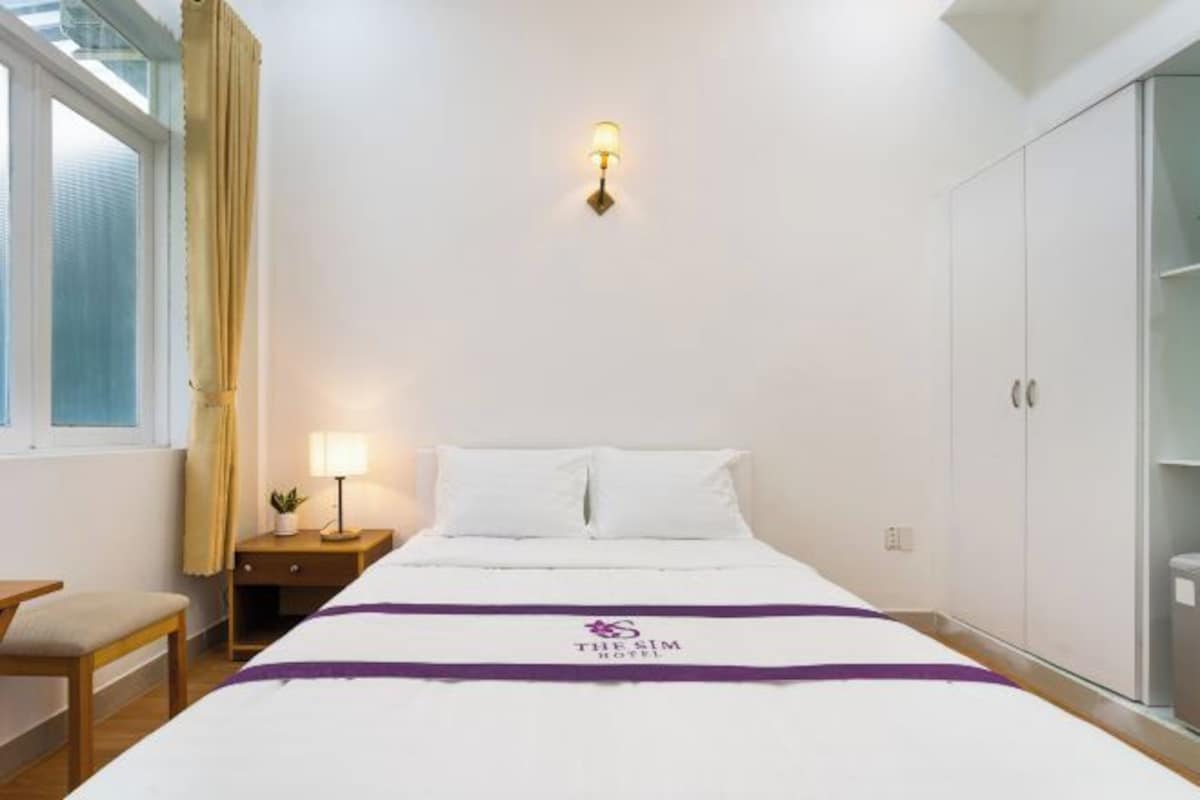 A hotel room with a bed, a nightstand with a lamp, a window with curtains, and a wardrobe.