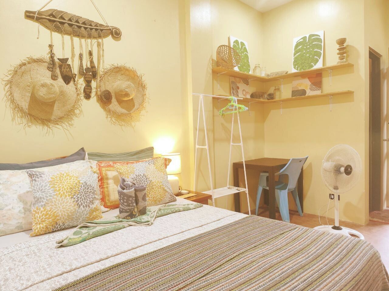 Un dormitorio ordenado con cama, mesitas de noche, un escritorio y un ventilador.