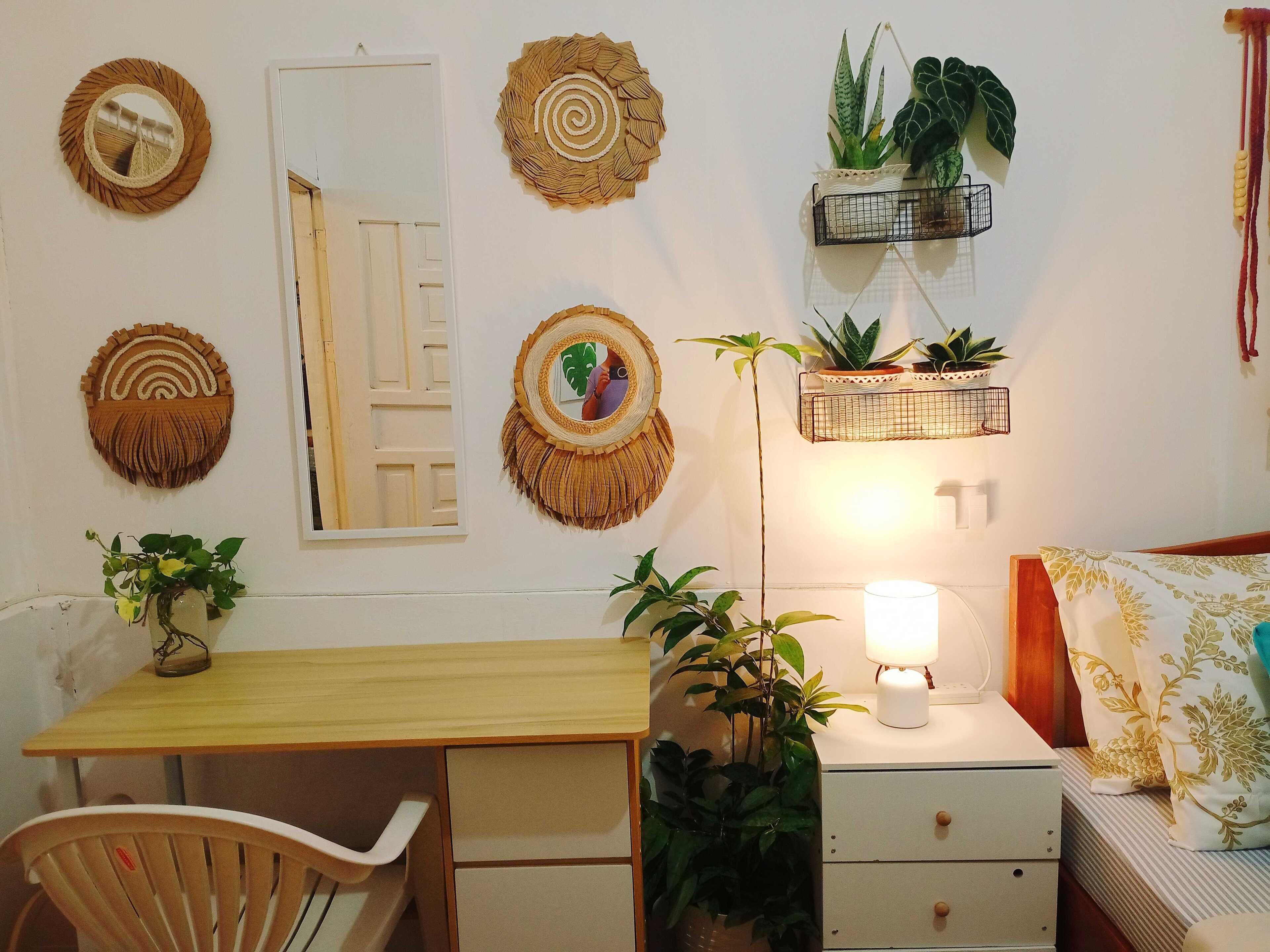 A room with a wooden desk, a chair, a bedside table with a lamp, a bed with patterned bedding, and wall-mounted decor including woven wall hangings and potted plants.