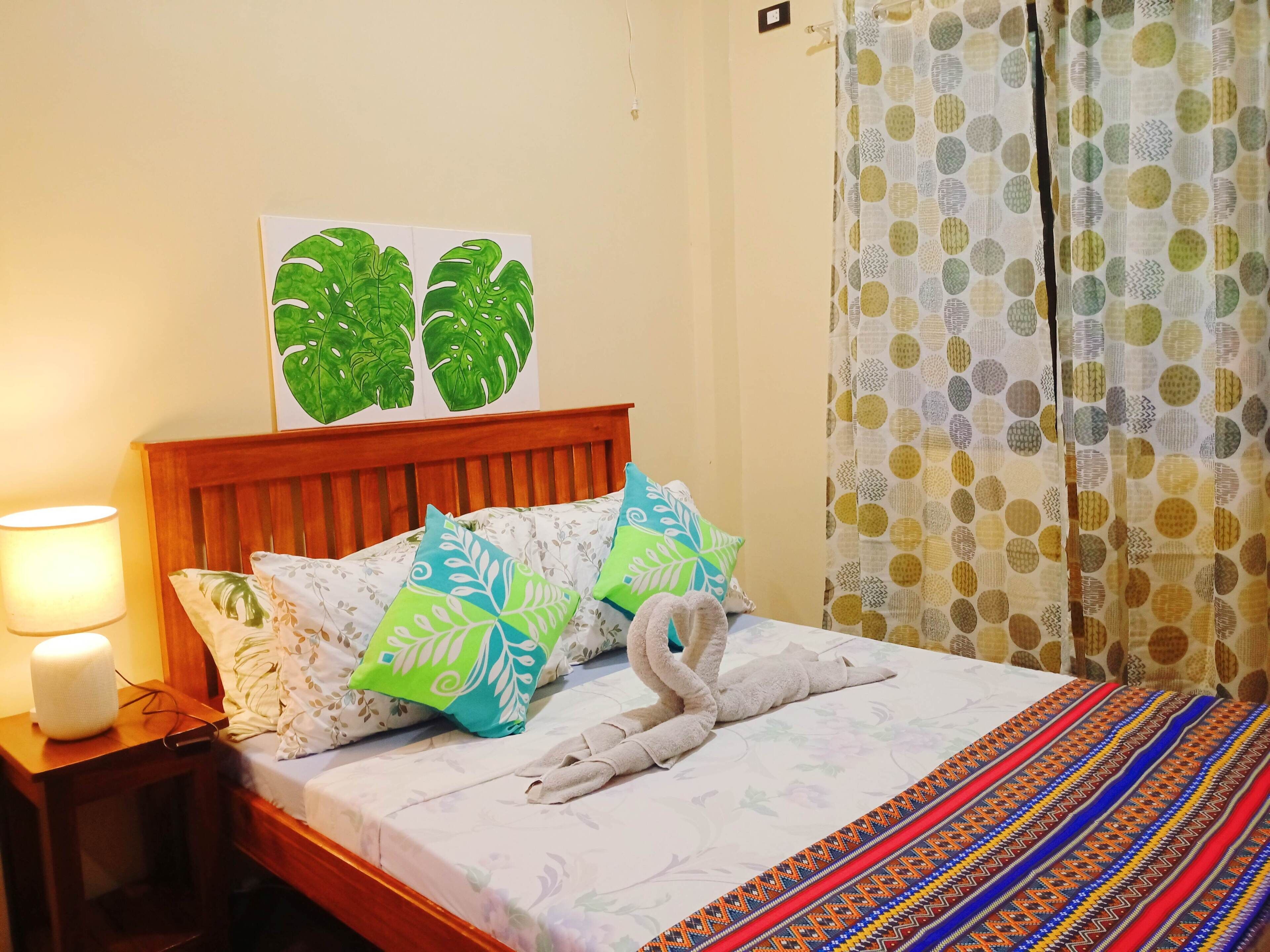 A neatly made bed with a floral-patterned bedspread, a decorative swan towel, and a pair of green and blue pillows.