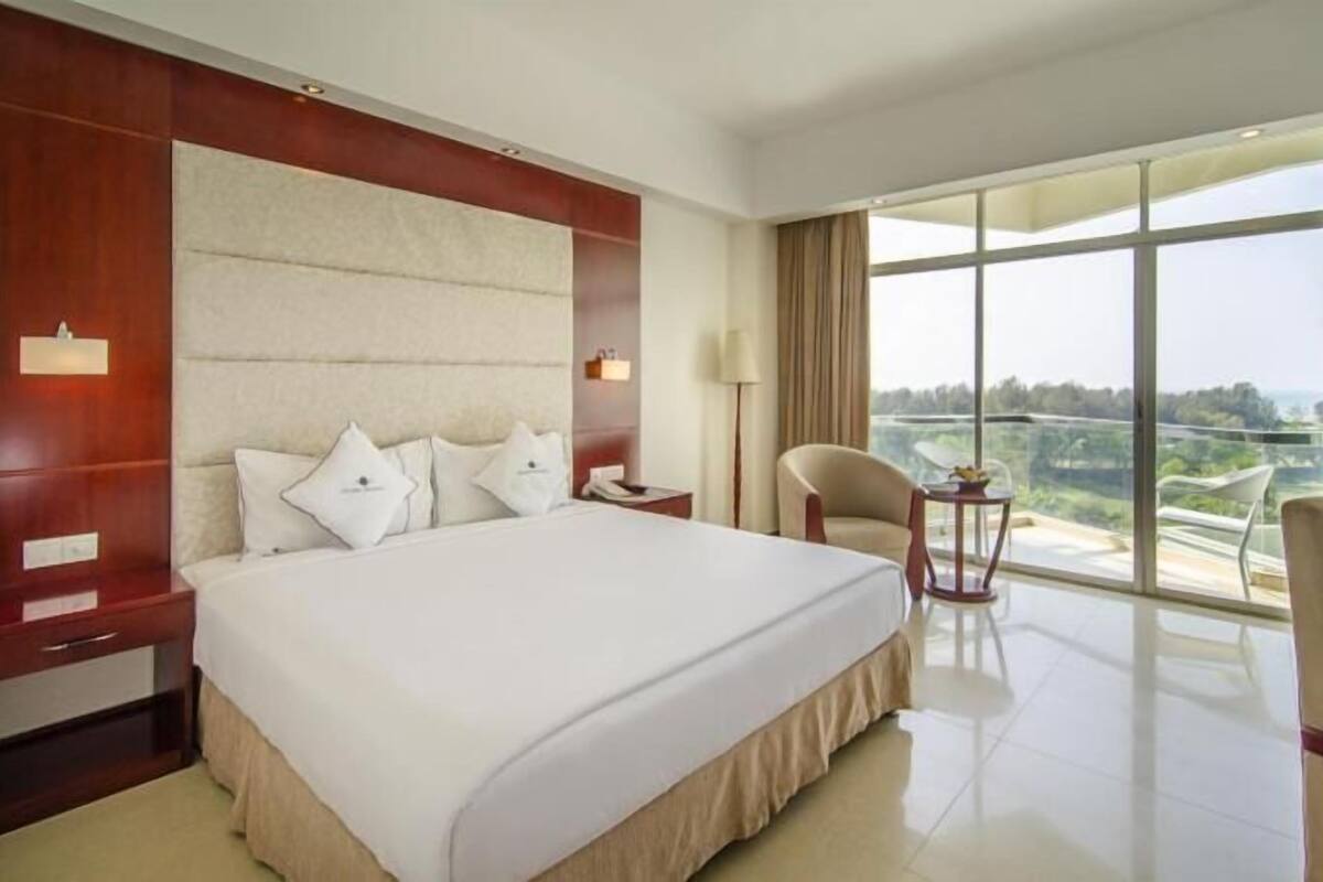 A large bed with a headboard, a bedside table, a chair, and a view of the outdoors.