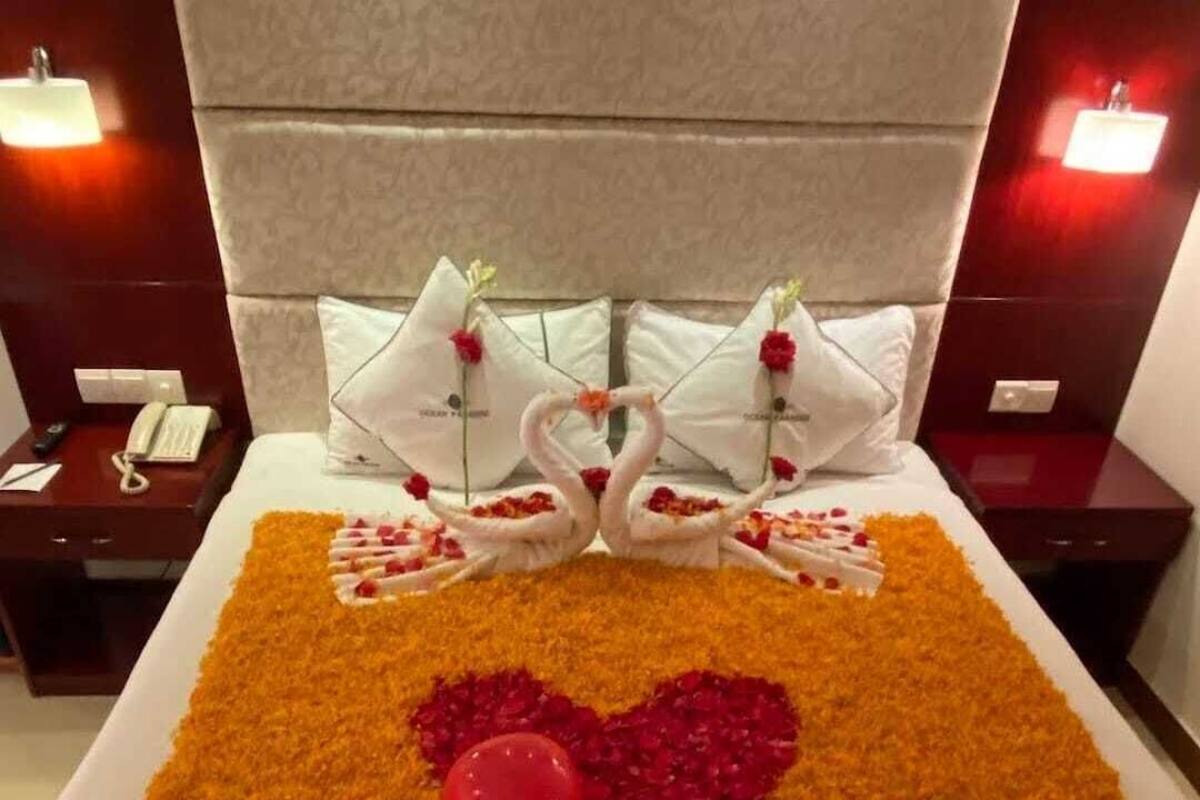 A hotel room with a bed covered in orange petals forming a heart shape, two swan-shaped pillows with flowers, and a red rose on the bed.