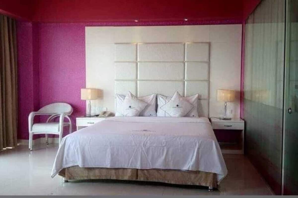 A bedroom with a large bed, a white chair, a bedside table with a lamp, and a glass shower enclosure.