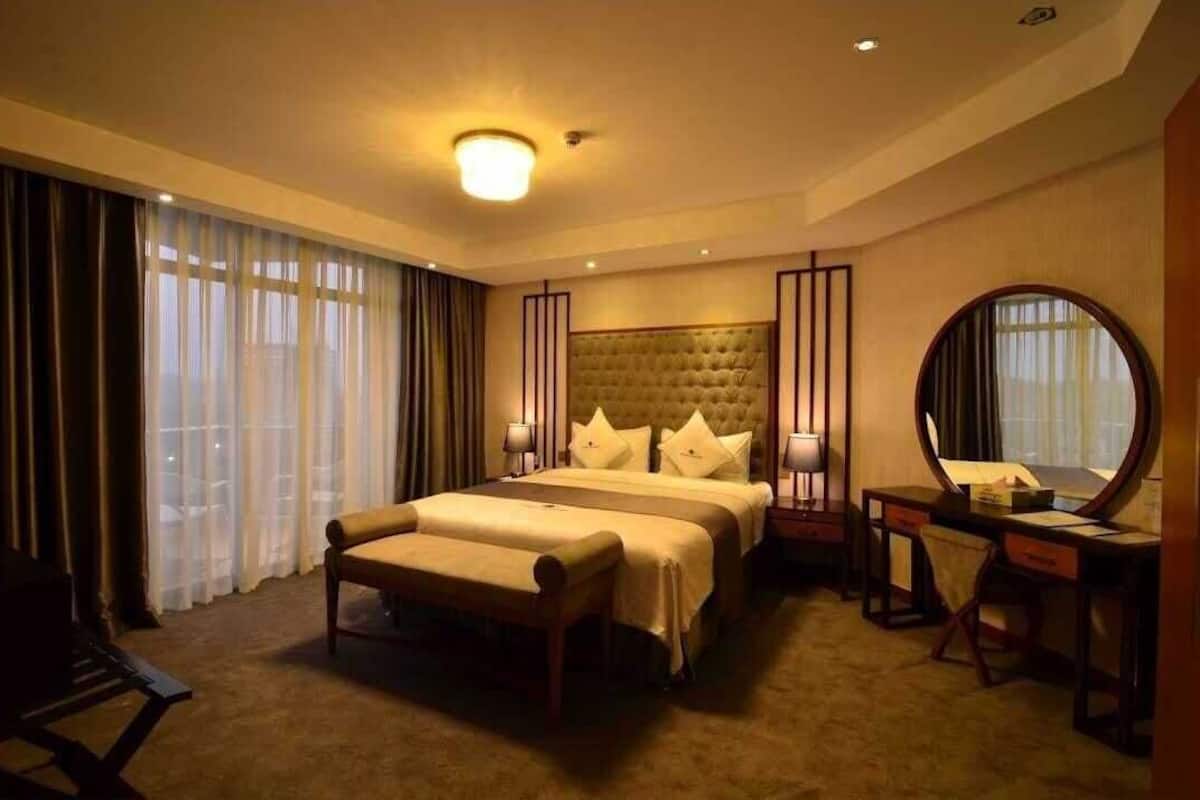 A hotel room with a large bed, bedside tables, a desk with a chair, a round mirror, and a large window with curtains.
