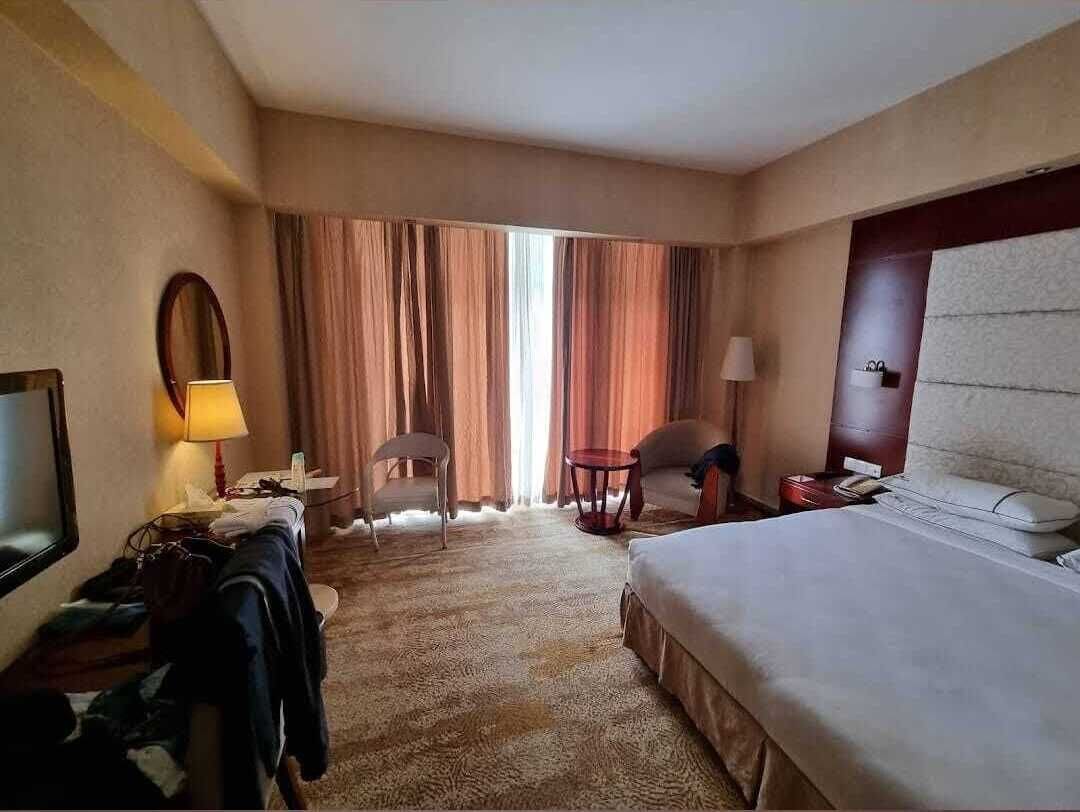 Une chambre d’hôtel avec un lit, un bureau doté d’une lampe, une chaise, une table ronde et une grande fenêtre avec des rideaux.
