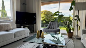 A modern living room with a sofa, a glass coffee table, a television, and a large window with a view of trees and a lawn.