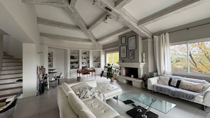 A spacious living room with a high ceiling, exposed beams, a fireplace, and a large window with a view of trees.