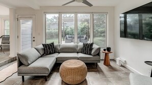 Un salon moderne avec un canapé d’angle gris, un pouf rond en osier, un ventilateur de plafond et un grand téléviseur fixé au mur.