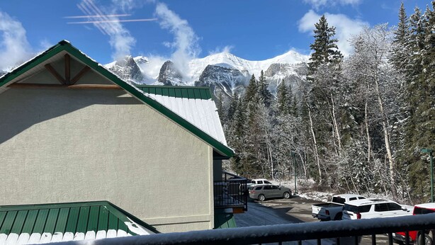A building with a green roof, a parking lot with several cars, and snow-covered trees and mountains in the background. - Banff Woods Lodge (Harvie Heights)