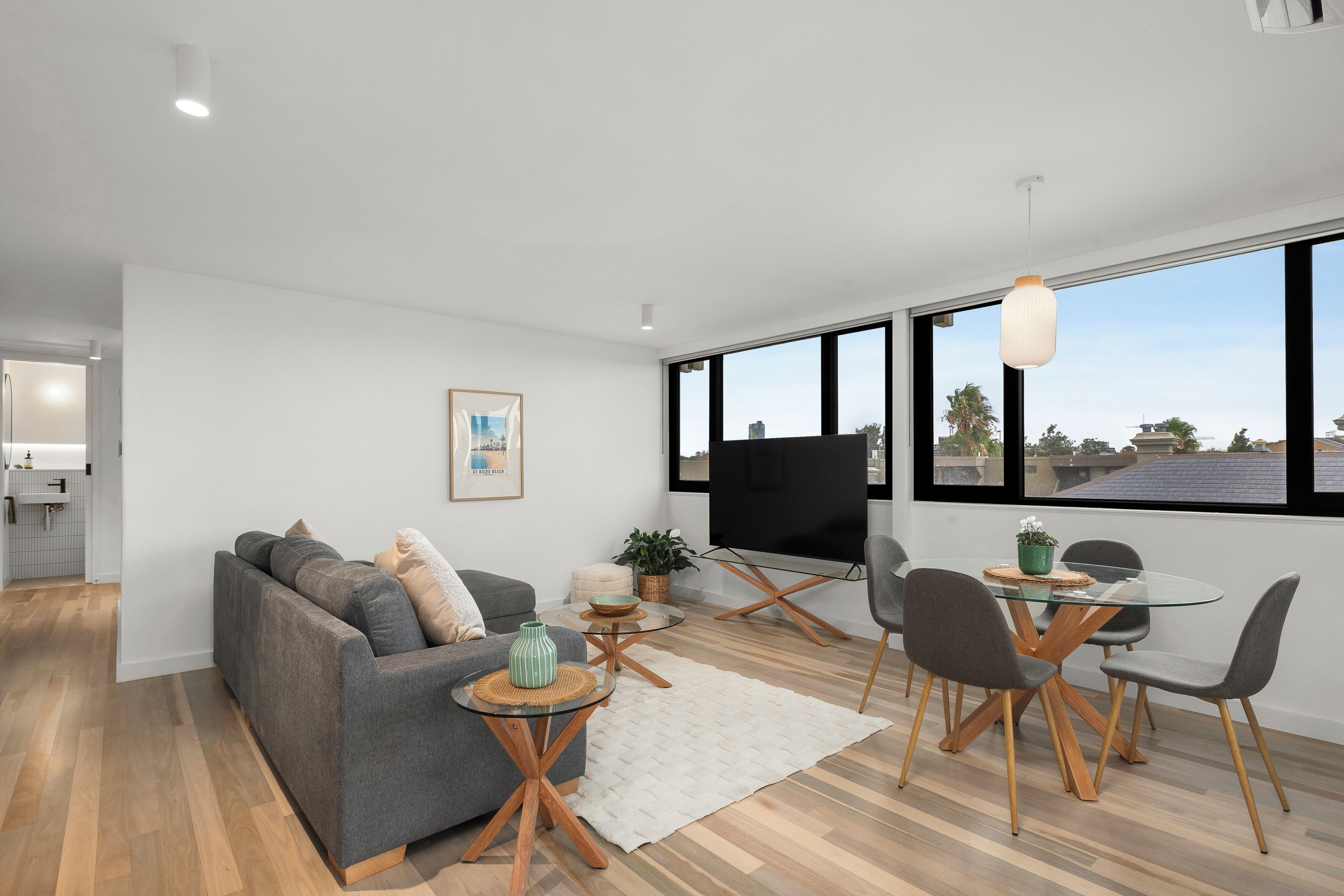 A modern living room with a grey sofa, a glass dining table with chairs, a TV, and a large window with a view of the city.