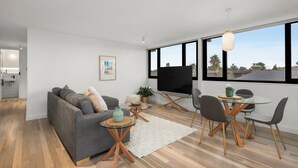 Ein modernes Wohnzimmer mit grauem Sofa, einem Glastisch mit Stühlen, einem Fernseher und einem großen Fenster mit Blick auf die Stadt.