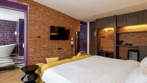 A modern hotel room with a brick wall, a large bed, a bathtub, and a flat-screen TV mounted on the wall.
