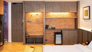 A modern bedroom with a brick wall, wooden furniture, a bed, a small seating area, and a mini-fridge.