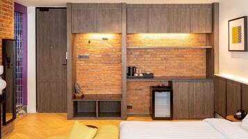 A modern bedroom with a brick wall, wooden furniture, a bed, a small seating area, and a mini-fridge.
