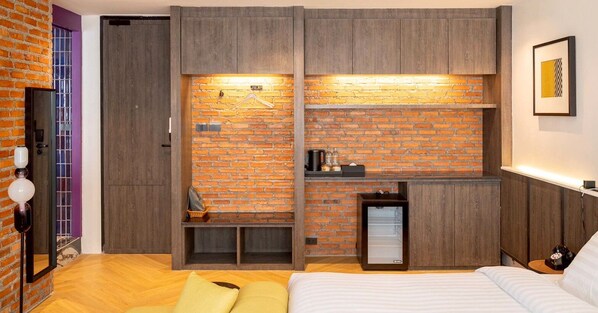 A modern bedroom with a brick wall, wooden furniture, a bed, a small seating area, and a mini-fridge. - Santisookmidtown (Pattani)