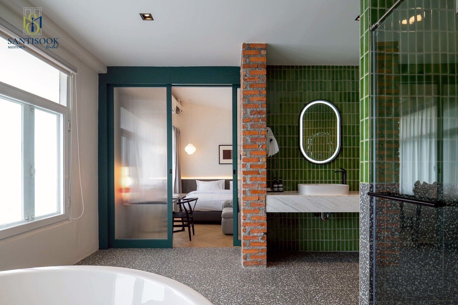 Une salle de bain moderne avec une baignoire, un lavabo et un miroir. Une porte donne sur une chambre à coucher équipée d’un lit et d’une chaise. La salle de bain est décorée de carreaux verts et possède un mur d’accent en briques.