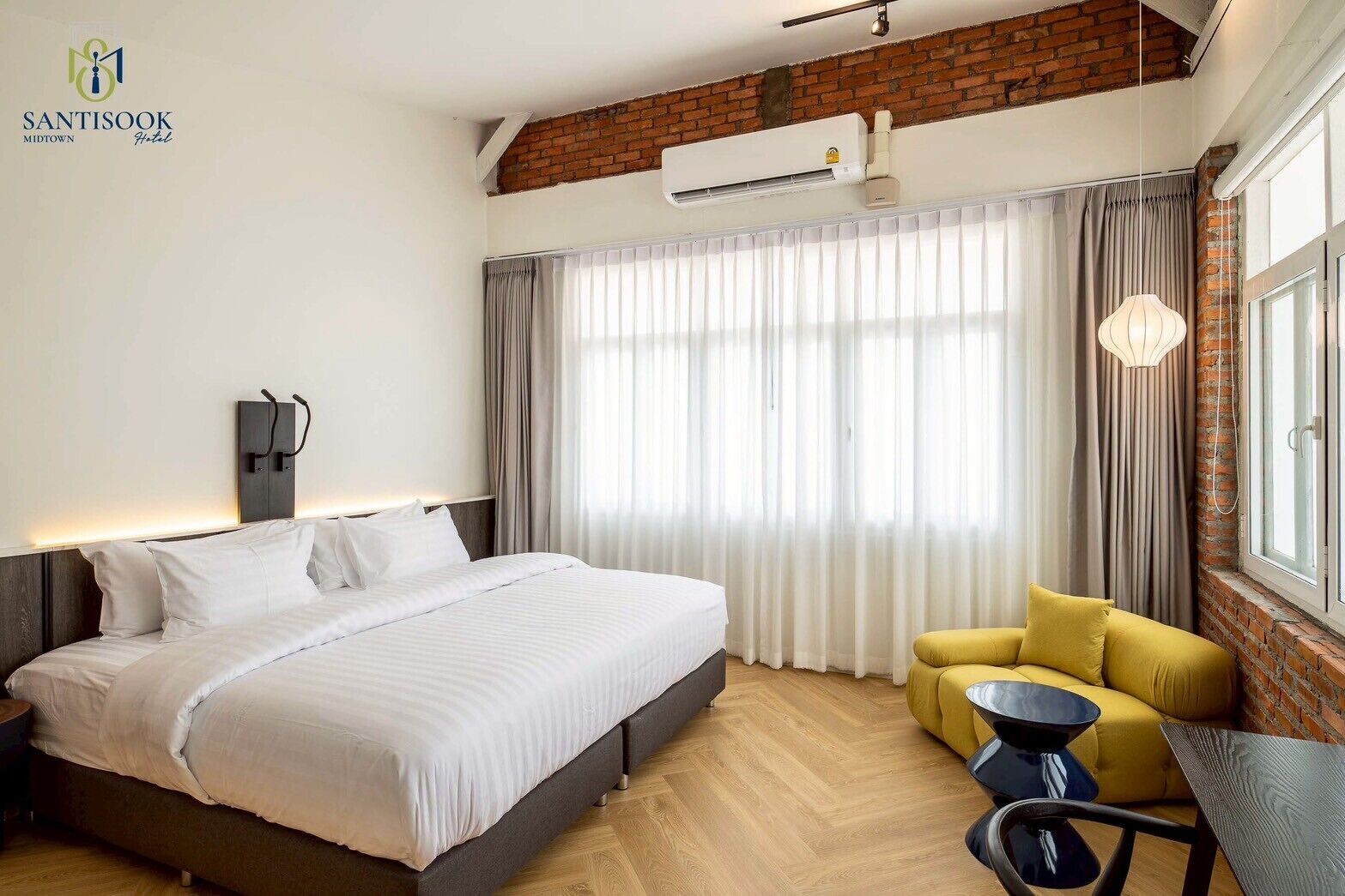 Une chambre d’hôtel avec un lit, un fauteuil jaune, une petite table et une grande fenêtre avec des rideaux.