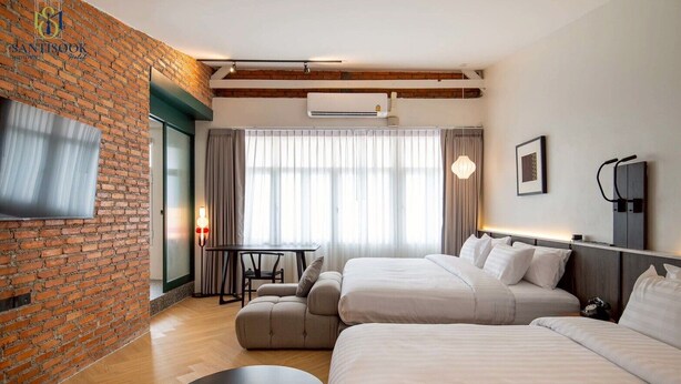 A hotel room with two beds, a TV, a brick wall, a window with curtains, and a small table with a chair. - Santisookmidtown (Pattani)