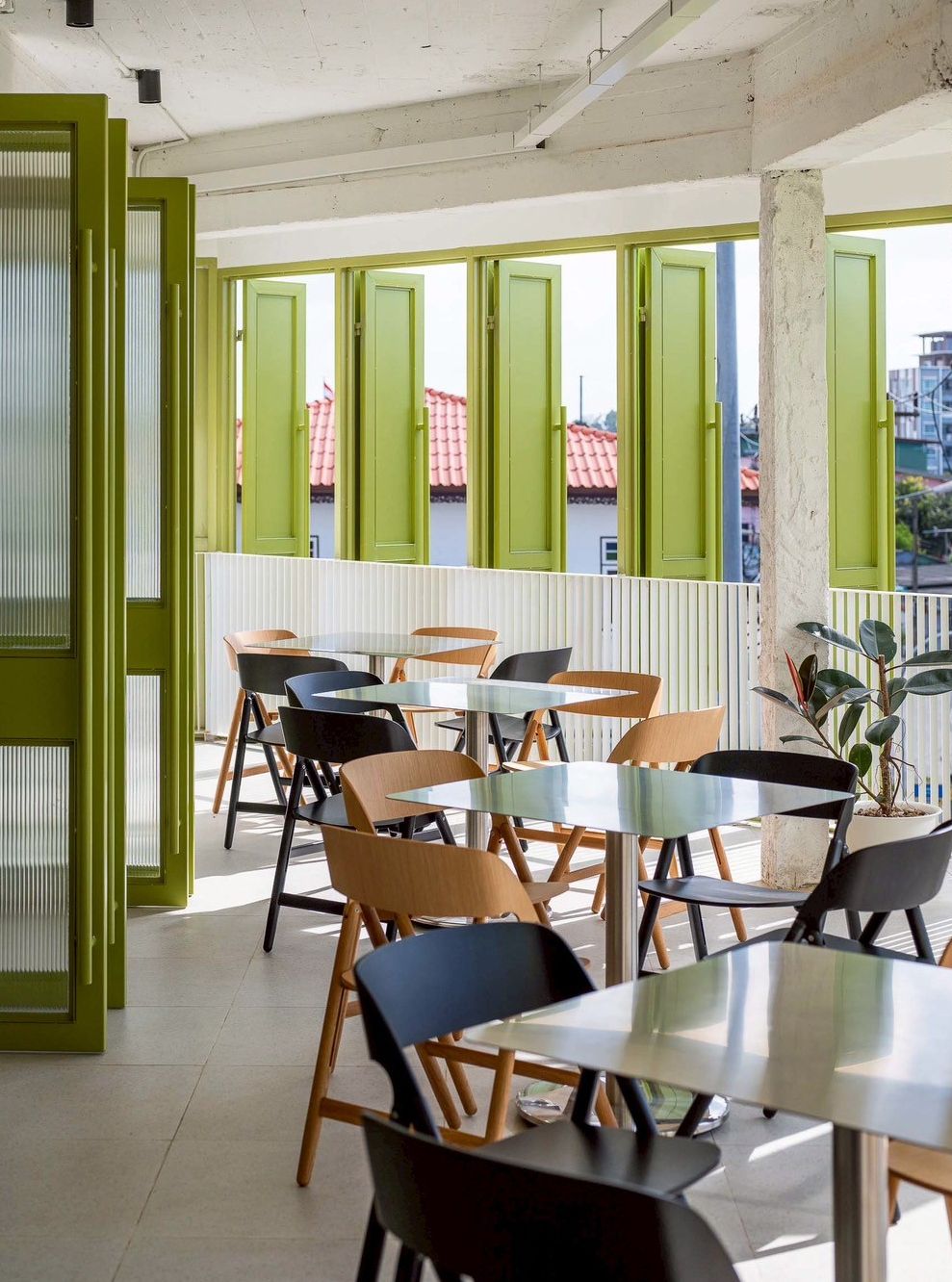 A dining area with tables and chairs, large windows with green shutters, and a view of buildings with red roofs.