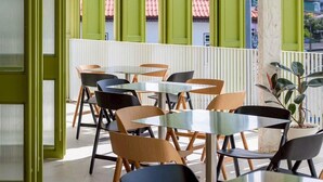 A dining area with tables and chairs, large windows with green shutters, and a view of buildings with red roofs. - Santisookmidtown (Pattani)