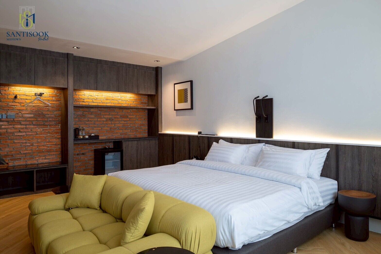 A modern hotel room with a large bed, a brick accent wall, a wooden nightstand, and a yellow armchair.