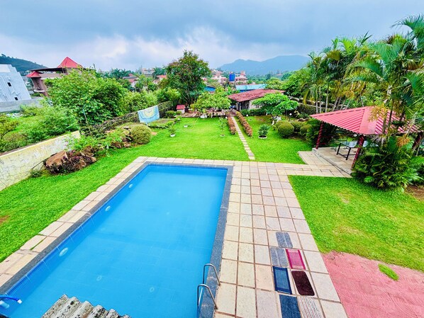 Une piscine dotée d’une terrasse carrelée, entourée d’une végétation luxuriante et offrant une vue sur des maisons et des montagnes situées au loin.