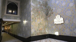 Un corridoio con parete piastrellata decorata con un motivo di alberi e frutta, una segnaletica per l'hotel Tillokori e un lucernario sopra.