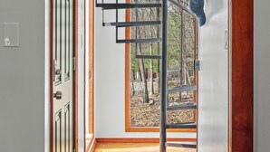 A modern interior with a spiral staircase, wooden flooring, and a view of a forest through a large window. - Lake Access + Pools + Sports Complex | Near Skiing & Hiking | Foxden (East Stroudsburg)