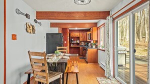 A dining area with a wooden table and chairs, a kitchen with wooden cabinets, and a view of the outdoors through large sliding glass doors. - Lake Access + Pools + Sports Complex | Near Skiing & Hiking | Foxden (East Stroudsburg)