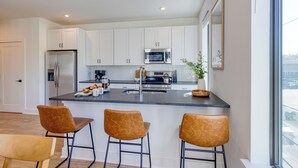A modern kitchen with white cabinets, a stainless steel refrigerator, a microwave, and a sink. There are three brown bar stools and a wooden dining table. - Stylish Condo • Private Balcony • Near Germantown + Broadway | The Heights (Nashville)