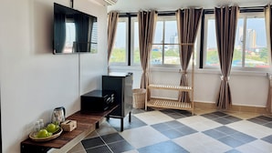 Quarto com televisão na parede, um armário preto, uma estante de madeira com utensílios de cozinha, uma janela com cortinas e vista para prédios.