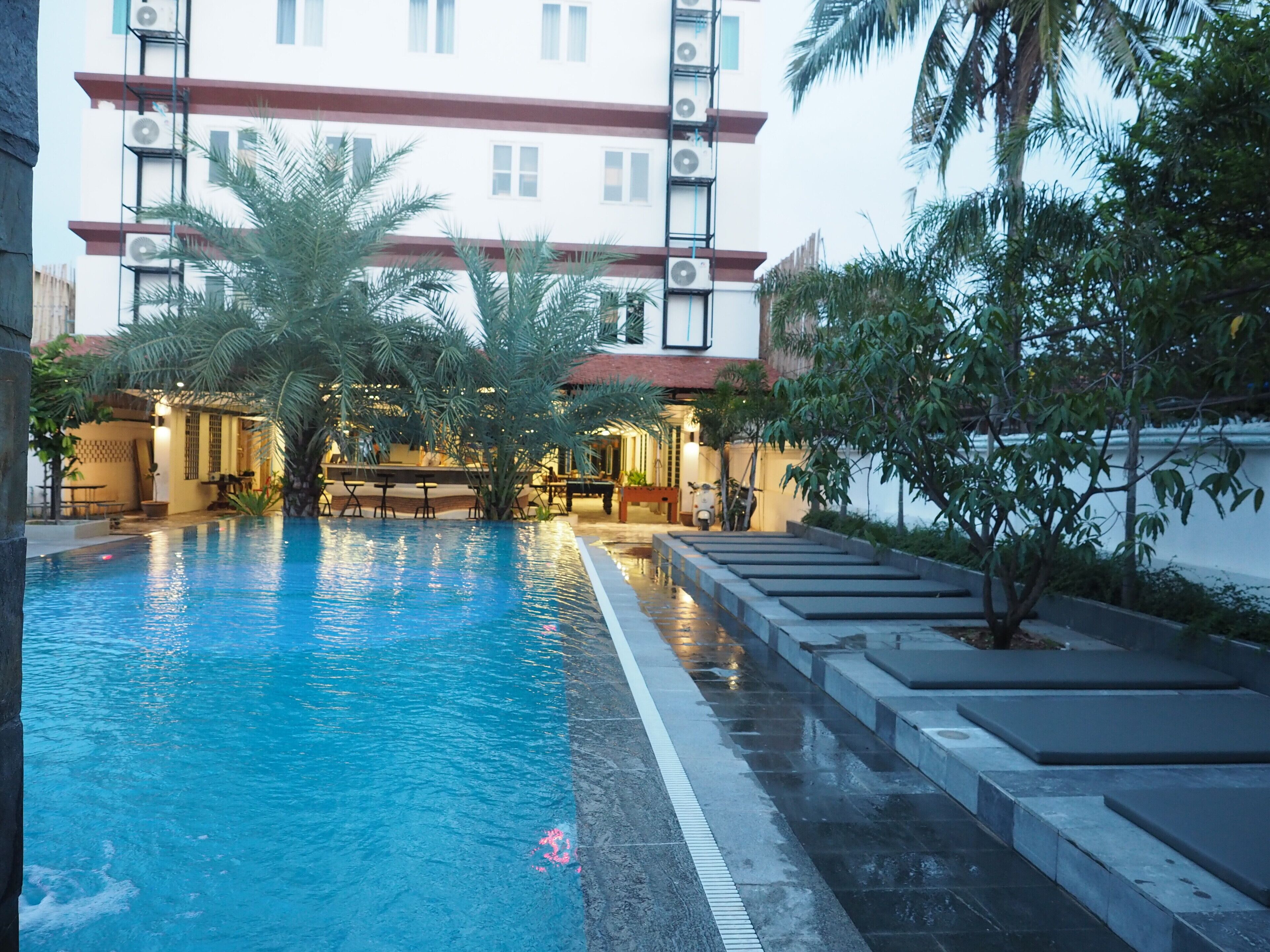 A swimming pool with steps leading into it, surrounded by palm trees and a building with air conditioning units.
