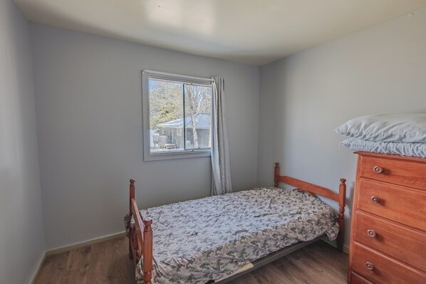 A small, single-bed room with a wooden headboard, a wooden dresser, and a window with curtains.