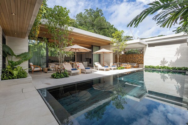 Un espace piscine extérieur moderne, doté de chaises longues et de parasols, entouré de verdure et agrémenté d’une terrasse en bois.