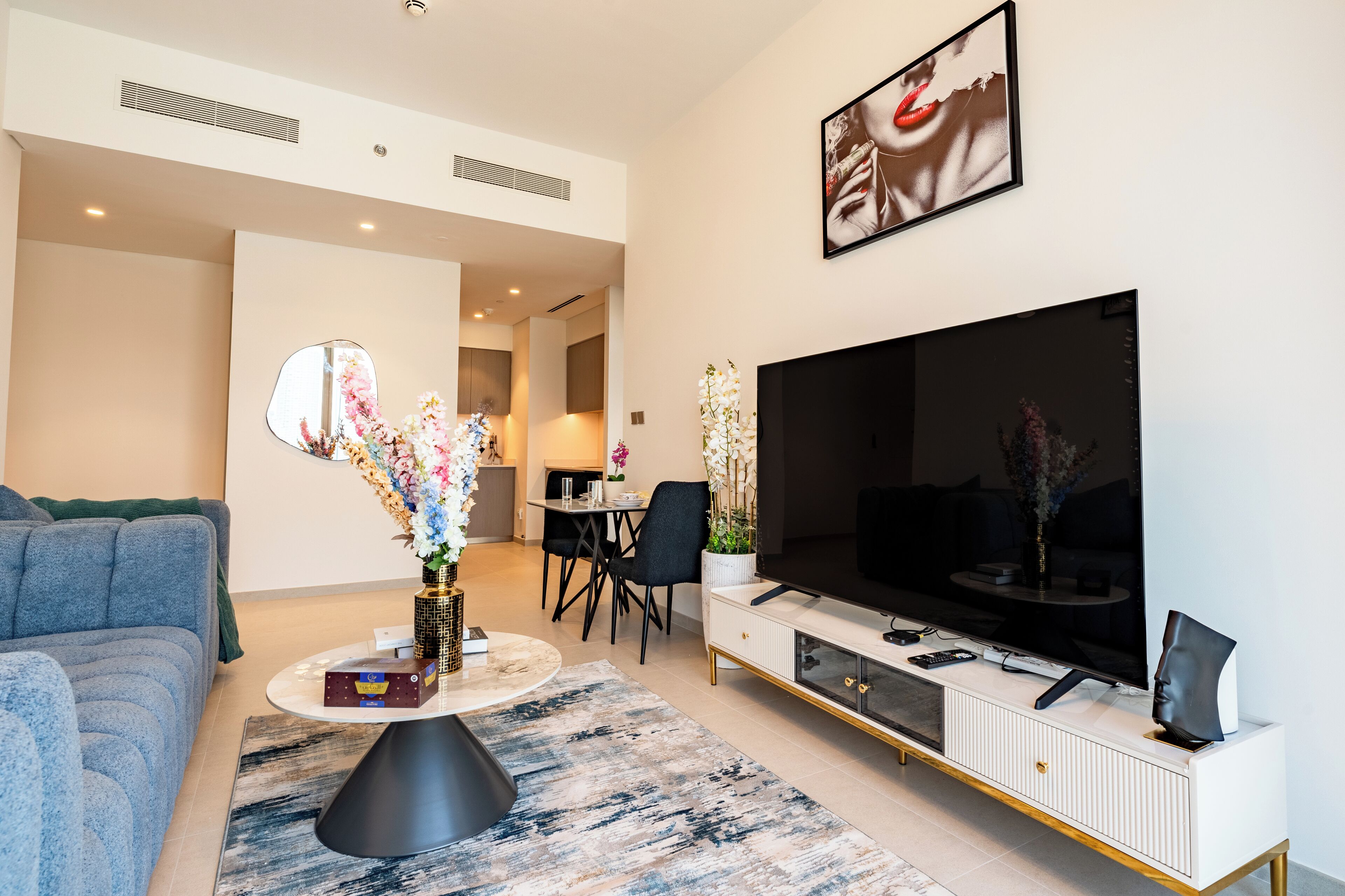 Ein modernes Wohnzimmer mit einer Couch, einem Couchtisch mit Blumenvase, einem Flachbildfernseher auf einer weißen Konsole und einem Essbereich im Hintergrund.