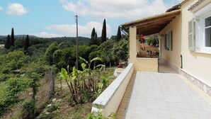 Una casa con un patio empedrado, un área de estar techada al aire libre y vistas a un paisaje verde con árboles y colinas.