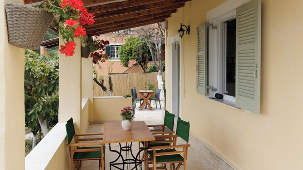 Un porche cubierto con una mesa y sillas de madera, una cesta colgante con flores y una ventana con contraventanas.
