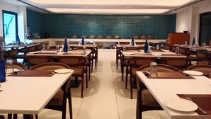 A dining area with multiple tables and chairs, a kitchen area with blue tiles, and a view of the outside through a window.