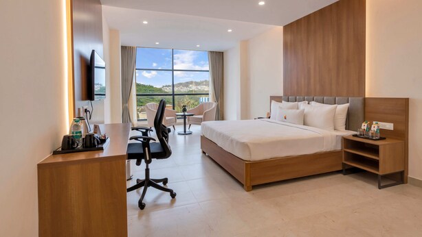 A modern hotel room with a large bed, a desk with office chair, a TV, and a view of the city.