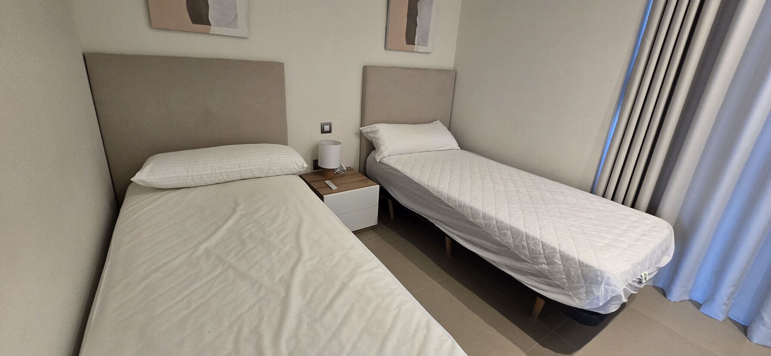 Une chambre d’hôtel avec deux lits simples, une table de chevet avec une lampe et une fenêtre avec des rideaux.