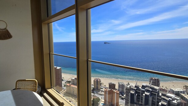 A room with a large window overlooking a beach and ocean. There are woven wall hangings and a wicker chair. - INTEMPO (2 BEDROOMS) (Benidorm)