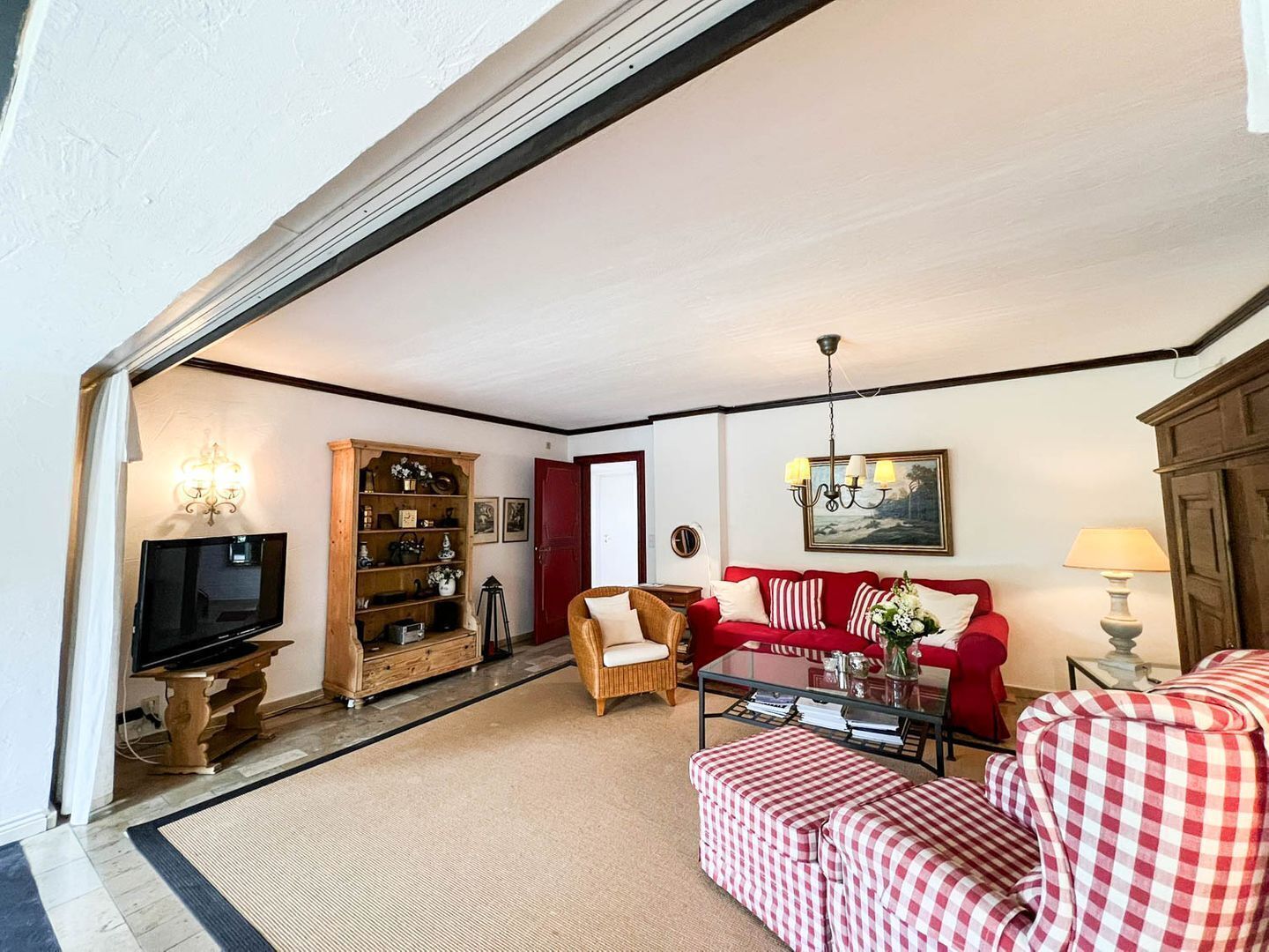 Una sala de estar con televisor, un sofá rojo, una silla de mimbre, un reposapiés a cuadros y una estantería.