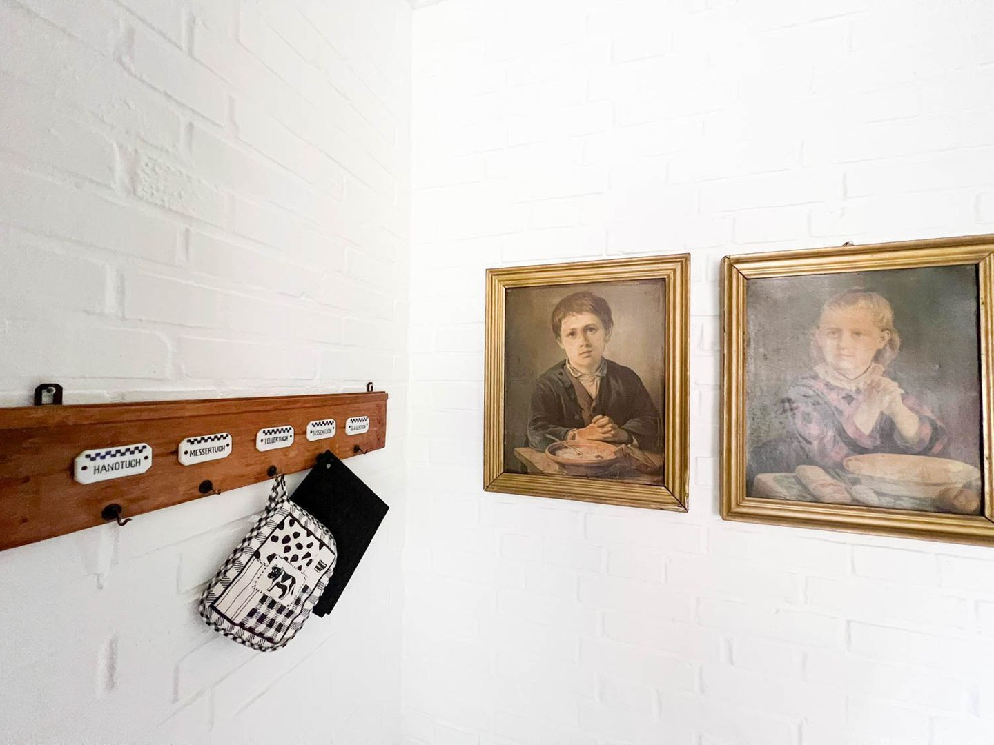 Pared de ladrillo blanco con dos retratos enmarcados y un estante de madera con objetos colgantes.