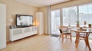 A modern living room with a flat-screen TV on a white cabinet, a dining area with wicker chairs, and large windows with sheer curtains.