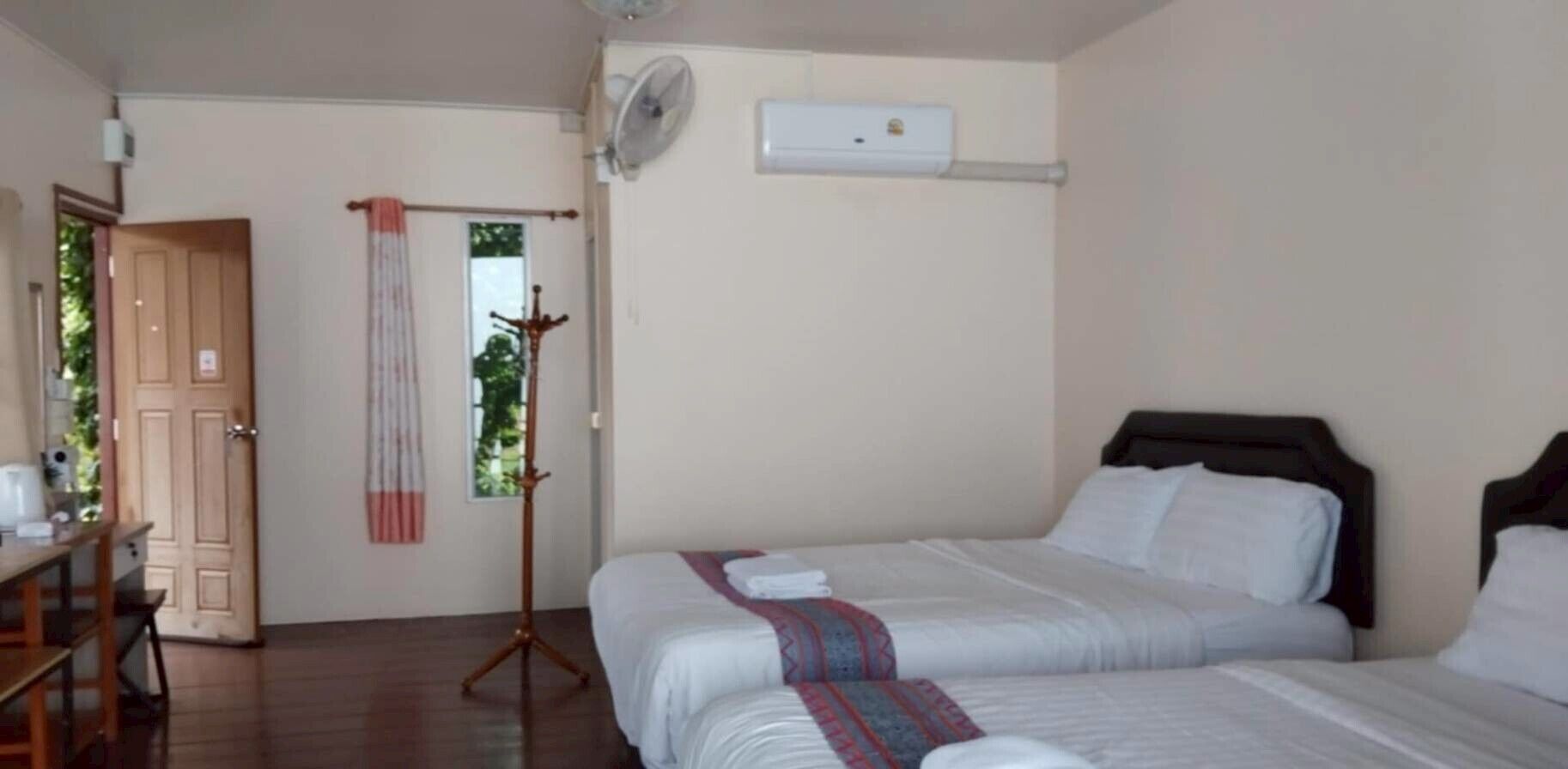 Habitación de hotel con dos camas, un escritorio de madera, un ventilador de pie y una ventana con vistas a follaje.