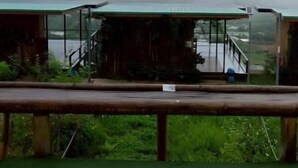 Una cabaña rústica con techo verde, una terraza de madera y vistas a montañas cubiertas de niebla.
