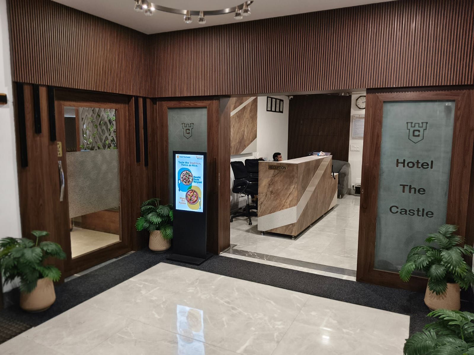 A hotel reception area with a reception desk, a digital screen displaying information, and a sign indicating the hotel name.